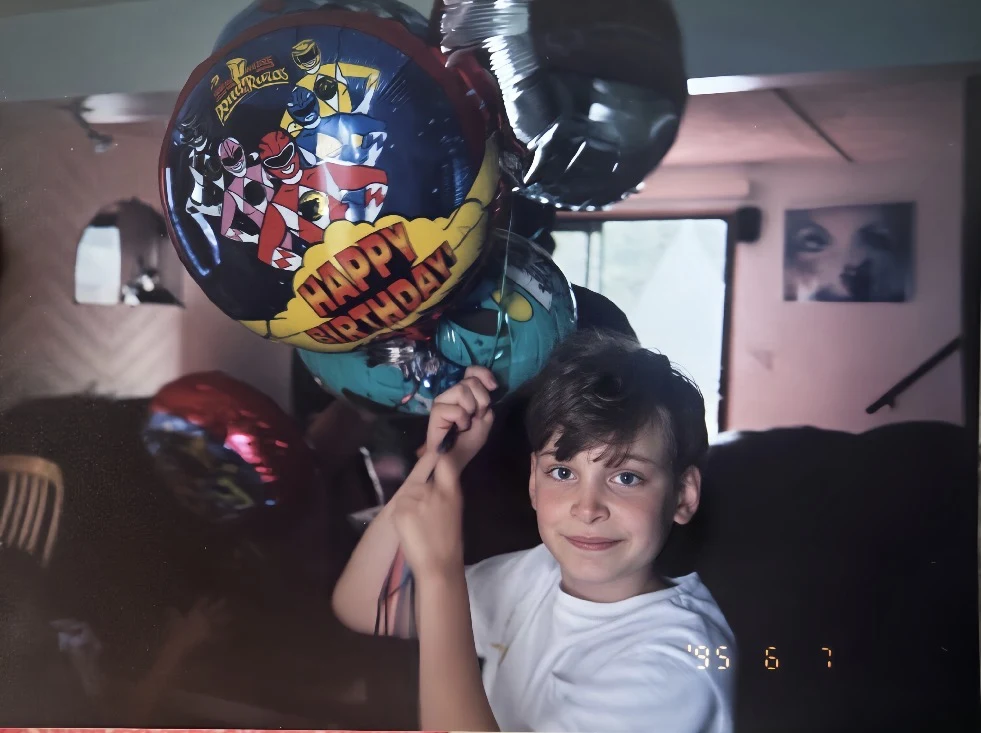 Young Erik Fuhrer at a birthday party in 1995, holding Power Rangers balloons. Hero image for essay on writing as survival and the final girl.